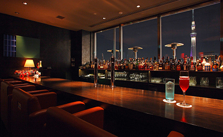 窓の外には紫に光る東京スカイツリーがあり、カウンターには赤と青のカクテル。奥にはたくさんのお酒が並び、オレンジのライトに包まれた落ち着ける雰囲気のバーの写真