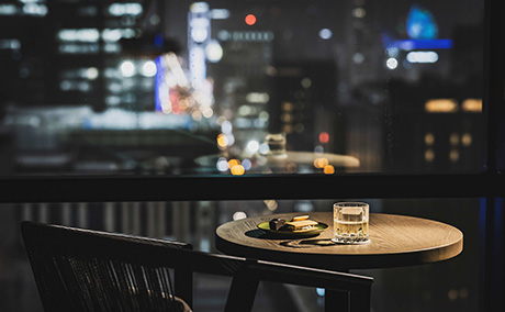夜の札幌を背景に、小さな丸いテーブルの上にお酒とスイーツがある洗練された大人の雰囲気漂うプレミアムラウンジの写真